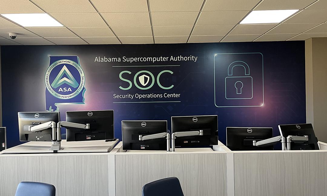 View of security operations center with rows of workstations in front of a large mural depicting the ASA logo, a SOC logo, and stylized lock icon